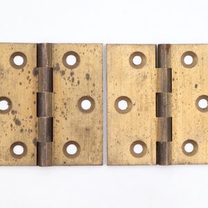 May include: Two vintage brass hinges with four screw holes each. The hinges are rectangular with a tarnished, aged appearance. The text "Belfast" and "Made in England" are visible on one hinge.