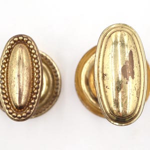 May include: Two antique brass door knobs. One knob has a beaded oval center, the other has a smooth oval center. Both knobs have a circular base. The brass has a worn, aged appearance.