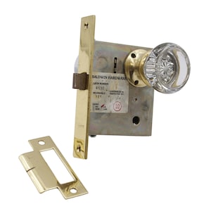 May include: A brass door knob set with a clear glass knob. The knob is a traditional style with a starburst pattern. The set includes a latch and strike plate. The label on the latch reads "Baldwin Hardware, Lock Number: R530, Reversible: YES, Assembled & Inspected By: 10".