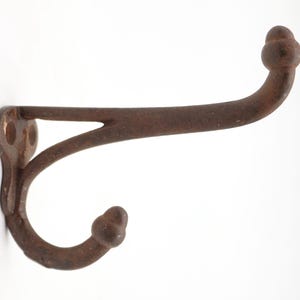 May include: A rustic, cast iron wall hook with a distressed, brown finish. The hook features a curved design with decorative ball-shaped ends. It has a mounting plate with two holes for easy installation. Ideal for hanging coats, hats, or other items.