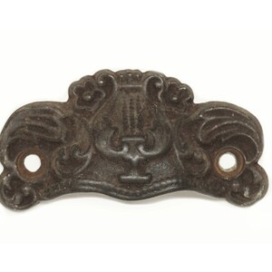 May include: A dark brown metal drawer pull with a decorative design featuring a lyre and floral motifs. The pull has two holes for attaching screws.