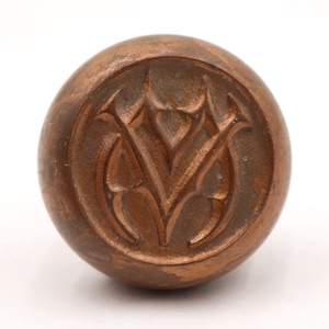 May include: A round, bronze-colored knob with a carved floral design. The knob has a smooth, polished surface and is likely made of metal. The design is centered on the knob and is a series of stylized leaves or petals.