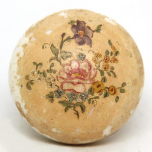 May include: A round, off-white decorative object with a floral design. The hand-painted flowers include a large pink rose, purple flower, and smaller yellow and blue blossoms. The object has a slightly aged appearance.
