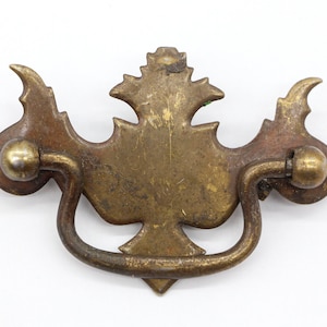 May include: Antique brass cabinet pull with a decorative, ornate design. The pull has a loop handle and a central, leaf-shaped plate.