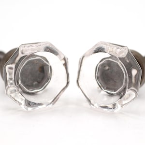 Op de afbeelding: Twee achthoekige kastknoppen van helder glas met gefacetteerde randen. Elke knop heeft een donkere metalen basis in het midden en een montageschroef aan de achterkant, met een transparant ontwerp en een geometrische vorm.