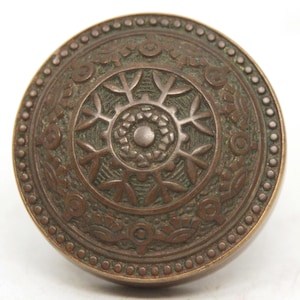 Puede incluir: Un pomo de puerta redondo de bronce antiguo con un diseño en relieve ornamentado. El pomo presenta un motivo floral central rodeado por un borde detallado de formas y puntos repetidos. El metal envejecido tiene una cálida pátina marrón.