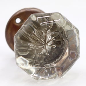 May include: A clear, octagonal glass doorknob with a starburst design is shown in a close-up shot. The knob is attached to a round, bronze-colored metal plate. The glass knob has a vintage aesthetic.