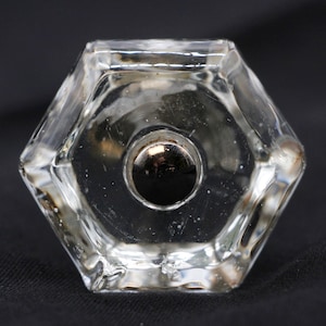 Puede incluir: Un pomo de puerta de vidrio transparente con forma hexagonal y un inserto de metal redondo en el centro.