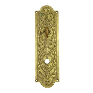 May include: Ornate brass door handle with a detailed, symmetrical design. The handle features a central pull and intricate floral and scrollwork patterns. The rectangular backplate has a keyhole and mounting holes.