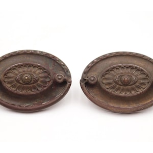 Puede incluir: Dos tiradores de cajón ovalados de bronce antiguo. Cada tirador presenta un diseño floral central y pequeños acentos circulares. Los tiradores tienen un acabado envejecido, lo que sugiere edad y uso. Ideal para proyectos de restauración de muebles antiguos.