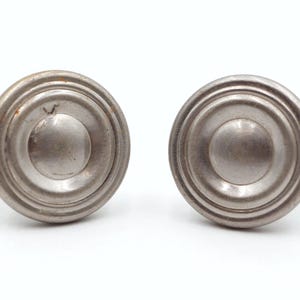 Peut inclure: Deux boutons de meuble anciens de couleur argentée. Chaque bouton présente un design circulaire avec des anneaux concentriques et un bouton central arrondi. Les boutons sont posés sur un fond blanc.