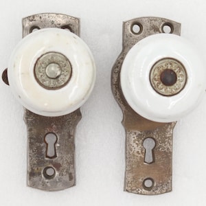 May include: Two vintage door knobs with white porcelain centers and weathered metal backplates. Each backplate features a keyhole cutout and two screw holes for mounting. The center of each knob has a decorative metal inlay with a circular pattern.