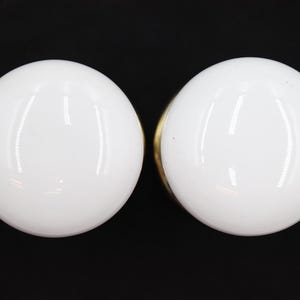 Könnte beinhalten: Zwei weiße, kugelförmige Schrankknöpfe mit glänzendem Finish. Die Knöpfe sind vor einem schwarzen Hintergrund platziert, der ihre glatten, reflektierenden Oberflächen hervorhebt. Die Hardware scheint neu und unbenutzt zu sein.