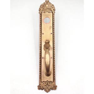 May include: Ornate antique door handle and backplate in a gold tone. The handle is a curved bar with a decorative backplate featuring intricate scrollwork and a circular opening. The design is symmetrical and classic.