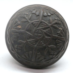 May include: A black, round, metal door knob with an intricate, carved design. The design features a central circle with radiating lines and a border of repeating floral motifs.