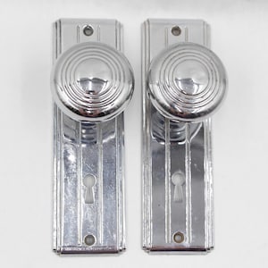 May include: Two vintage chrome door knobs with matching backplates. The knobs are round with concentric circles, and the backplates have vertical grooves. Each plate has a keyhole and a small hole at the bottom. The hardware is likely from the Art Deco era.