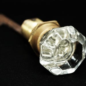 Pode incluir: Puxador de porta de vidro transparente e facetado com base de latão e haste marrom escuro. O puxador tem formato octogonal e reflete a luz, em fundo preto. Um item de hardware vintage.