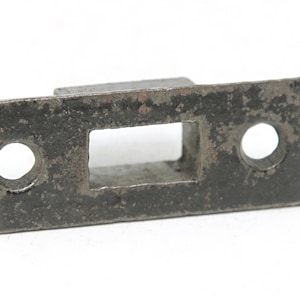 May include: A rectangular, weathered metal door strike plate with a square opening in the center and two circular holes. The plate has a dark, aged appearance with signs of rust.