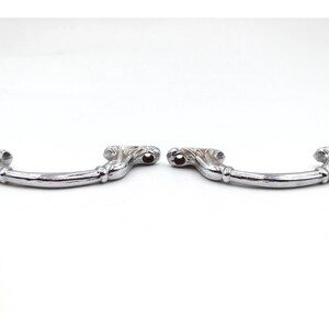 May include: Two silver-colored drawer pulls with a curved handle design. Each pull features ornate detailing on the ends and a smooth, polished finish. The pulls are designed for furniture and cabinet hardware.