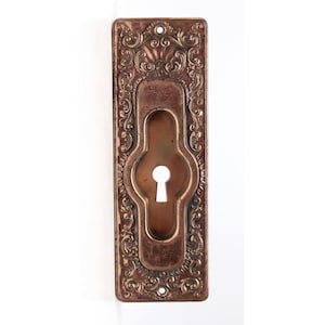 Puede incluir: Una cubierta de mirilla vintage con un diseño ornamentado. La cubierta está hecha de metal y tiene un acabado de cobre. Es de forma rectangular y tiene una mirilla en el centro.