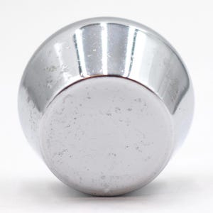 Pode incluir: Um objeto esférico cromado brilhante com uma base plana. A superfície reflete a luz, mostrando pequenas imperfeições. O objeto parece ser um elemento decorativo ou parte de um item maior.