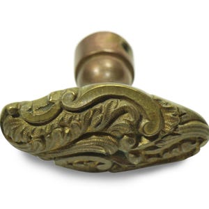 Könnte beinhalten: Ein antiker Türknauf aus Messing mit einem verzierten, floralen Design. Der Knauf hat eine abgerundete Oberseite und einen detaillierten, oval geformten Griff. Das Messing hat eine warme, gealterte Patina, die auf seinen Vintage-Ursprung hindeutet.