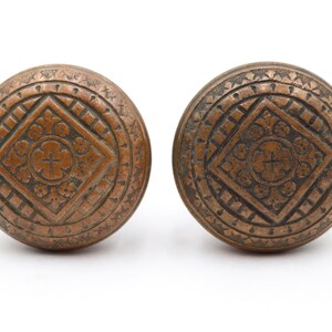 May include: Two antique bronze door knobs with intricate geometric and floral patterns. Each knob features a central diamond shape with a cross design, surrounded by detailed circular borders. The knobs are displayed against a white background.