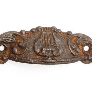 May include: A vintage cast iron drawer pull with an ornate design featuring a lyre, a musical instrument. The pull is a dark brown color with rust accents.