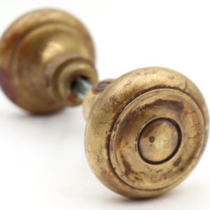 Peut inclure: Bouton de porte ancien en laiton avec une finition patinée. Le bouton a un design rond et étagé avec une indentation circulaire centrale. Le laiton a une teinte dorée chaude avec des zones de patine plus foncée, suggérant l'âge et l'utilisation. Le bouton est monté sur une tige métallique.