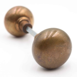 May include: A pair of antique brass door knobs. Each knob is a rounded sphere with a tarnished, aged appearance. The knobs are connected by a silver-colored metal rod, set against a white background.