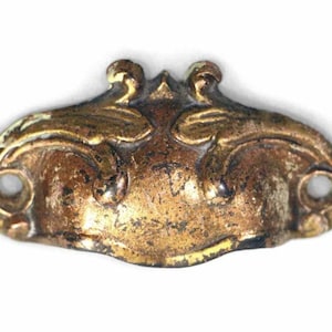 Może przedstawiać: Złoty metalowy uchwyt do szuflady z ozdobnym, bogatym wzorem. Uchwyt ma lekko zakrzywiony kształt i dwa otwory montażowe.