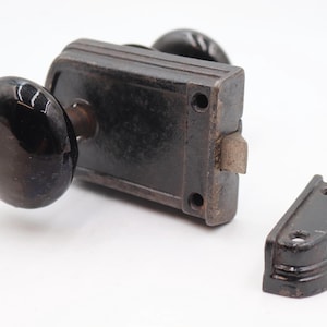 May include: An antique door lock set with a dark finish. The set includes a rectangular lock body, two round doorknobs, and a strike plate. The doorknobs are a glossy black color. The lock is likely made of metal.