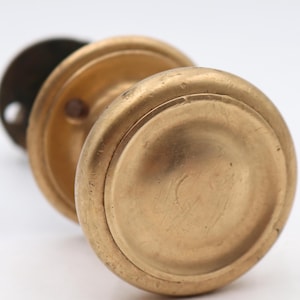 May include: Close-up of a vintage brass doorknob set against a white background. The doorknob has a round, aged appearance with a circular backplate. The brass has a warm, golden hue, suggesting age and use. The design is simple and classic.