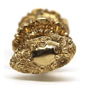 May include: A gold-coloured metal decorative knob with an ornate design. The knob has a rounded oval shape with a raised centre and intricate details.