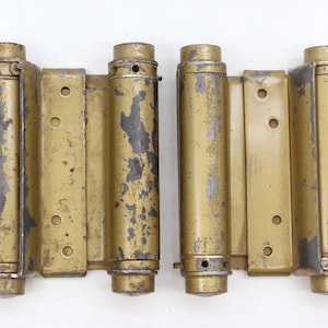 May include: Two gold-toned metal door hinges with multiple connecting pieces. The hinges are worn and have a distressed look.