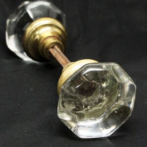 Puede incluir: Pomos de puerta de cristal transparente y facetado con herrajes de latón. Los pomos tienen una estética vintage y están conectados por una varilla metálica. El cristal es transparente y el latón tiene un tono cálido y envejecido.