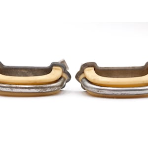 May include: Two antique drawer pulls. Each pull features a curved cream-coloured handle set within a metal frame. The metal has a distressed finish, giving them a vintage appearance. Ideal for furniture restoration.