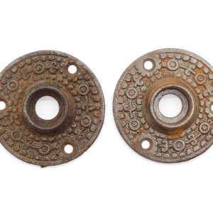 Może przedstawiać: Dwa antyczne, okrągłe gałki drzwiowe z żeliwa z misternymi wzorami. Gałki są rdzawo brązowe i mają wypukły środek z otworem pośrodku.