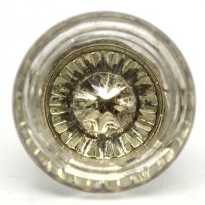 May include: A clear, antique-style glass doorknob with a ridged outer edge and a detailed, starburst-like center. The knob has a slightly aged appearance, suggesting vintage or retro design. The doorknob is a decorative hardware element.