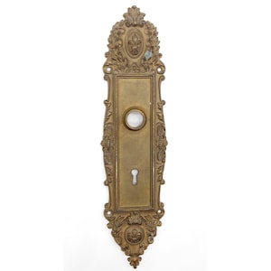 Op de afbeelding: Geornamenteerde goudkleurige metalen deurplaat met een sleutelgat en een ronde opening. De plaat heeft een decoratief ontwerp met bloem- en bladmotieven.