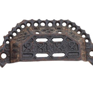 May include: A decorative, antique-style drawer pull made of dark brown metal. The pull has a semi-circular shape with intricate geometric and floral patterns, and small holes along the top edge. It features two holes for mounting.