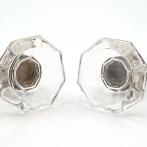 Op de afbeelding: Twee heldere glazen deurknoppen met een achthoekige vorm en een gefacetteerd oppervlak. Elke knop heeft een centrale metalen basis met een donkerbruine afwerking, met een geometrisch ontwerp dat verbonden is met een kleine, ronde steel.