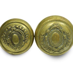 May include: A pair of antique brass door knobs. Each knob features a round shape with an oval central design, framed by decorative details. The brass has a warm, aged patina, indicative of vintage craftsmanship.