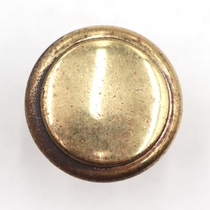 May include: A round, antique brass knob with a circular design. The knob has a central, slightly raised, brass-colored disc surrounded by a darker, aged brass rim. The surface shows signs of wear and age.