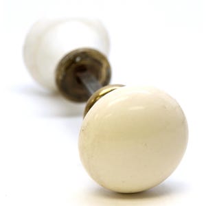 Peut inclure: Boutons de porte en céramique crème avec quincaillerie en laiton. Les boutons sont ronds et ont une finition brillante. La quincaillerie de connexion est en métal foncé. Le fond est blanc.