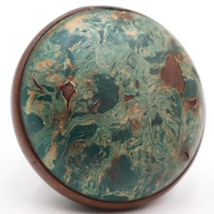 Op de afbeelding: Een ronde decoratieve knop met een gemarmerd groen en beige patroon. De knop heeft een donkerbruine houten rand en plekken met roodbruin materiaal. Het ontwerp geeft de knop een vintage of antieke uitstraling.
