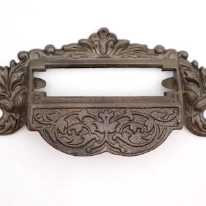Könnte beinhalten: Verzierte Schubladengriff in antikem Stil mit rechteckigem Etikettenhalter. Die dunkelbronze farbene Hardware weist detaillierte Blumen- und Scrollarbeiten auf. Enthält zwei Befestigungslöcher an jedem Ende. Ein dekorativer Akzent für Möbel.