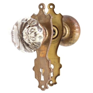 May include: Antique door hardware set featuring a clear, faceted glass doorknob and a brass backplate. The set includes a matching brass knob on the opposite side and a keyhole. The brass has a vintage patina.