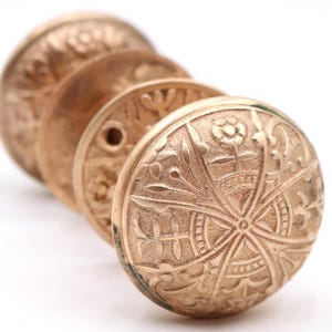 May include: A series of four round, metallic, brass-colored door knobs arranged in a line. Each knob features a detailed, embossed floral and geometric pattern on its circular face, with a smooth, rounded edge and a central stem extending backward.