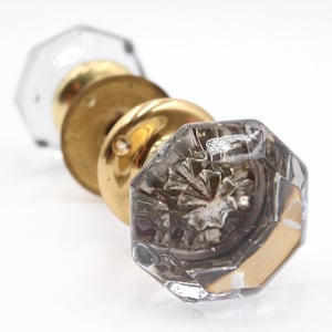 May include: Close-up of antique door knobs. The knobs feature a clear, faceted glass design with brass hardware. The glass has an octagonal shape. The brass hardware is circular and golden in color. The knobs are arranged in a row.